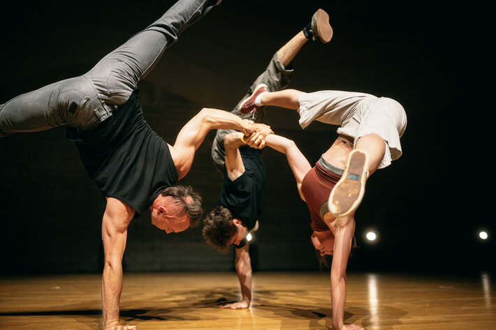 Drei Personen machen Akrobatik: Auf einer Hand stehend, die andere gegenseitig umfassend machen sie Handstand. (öffnet Vergrößerung des Bildes)