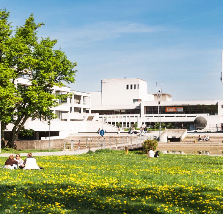 Campuswiese der Uni Regensburg mit Löwenzahn. Auf der Wiese sitzen Studierende. Im Hintergrund sieht man die Zentralbibliothek.
