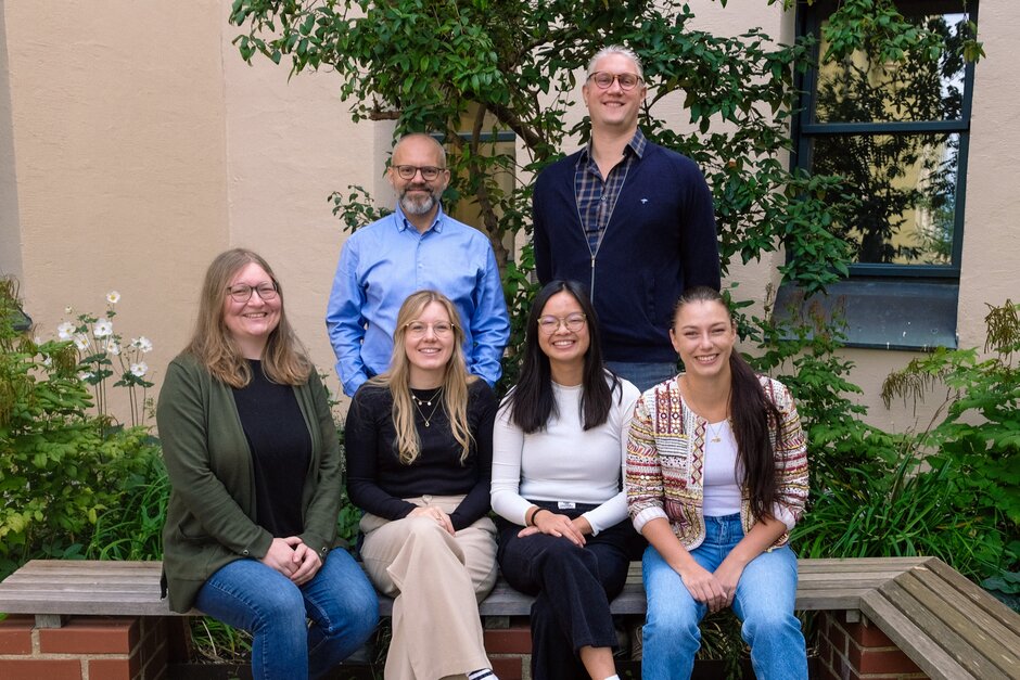 Vier Frauen sitzen auf Bank in Garten, dahinter stehen zwei Männer.