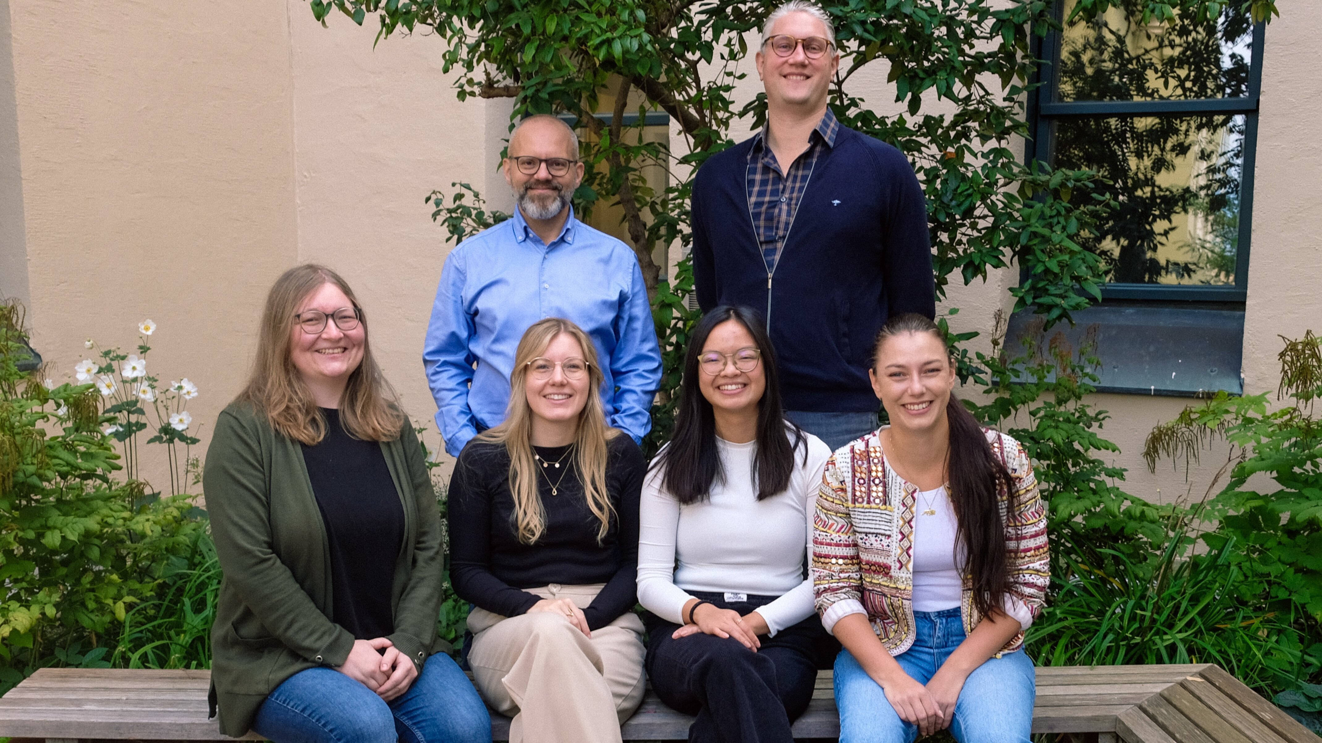 Vier Frauen sitzen auf Bank in Garten, dahinter stehen zwei Männer.