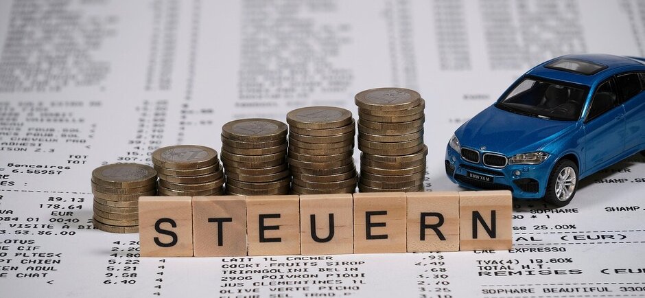 Ein Spielzeugauto und Stapel aus Geldmünzen, davor das Wort "Steuern" mit Buchstaben gelegt
