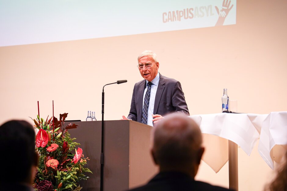 Mann an Rednerpult vor Mikrofon, im Vordergrund Publikum. (öffnet Vergrößerung des Bildes)