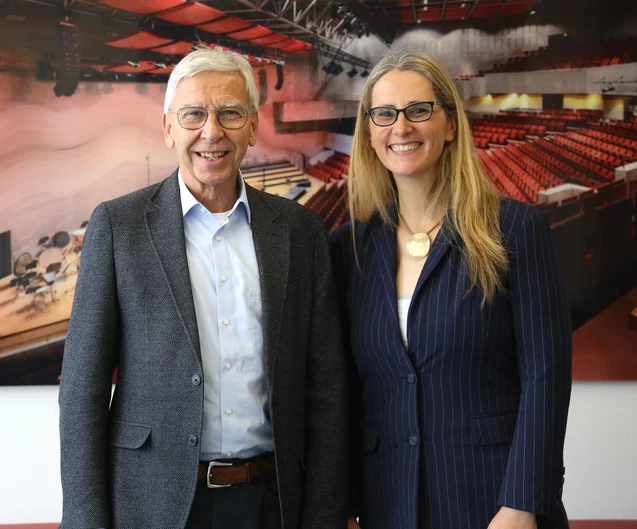 Ein älterer Mann und eine jüngere Frau stehen nebeneinander und lächeln in die Kamera. Beide tragen Brillen und formelle Kleidung. Im Hintergrund hängt ein großes Foto oder Gemälde eines Konzertsaals mit roten Sitzreihen an der Wand. Die Aufnahme scheint in einem Büro oder einem Veranstaltungsraum gemacht worden zu sein.