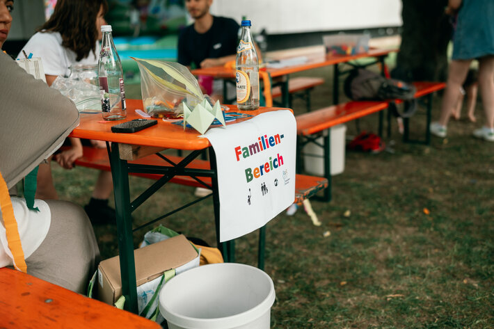 Das Bild zeigt einen Ausschnitt des Familien-Bereichs: Biertischgarnituren mit Beschriftung und im Hintergrund zwei sitzende Personen im Gespräch. (öffnet Vergrößerung des Bildes)