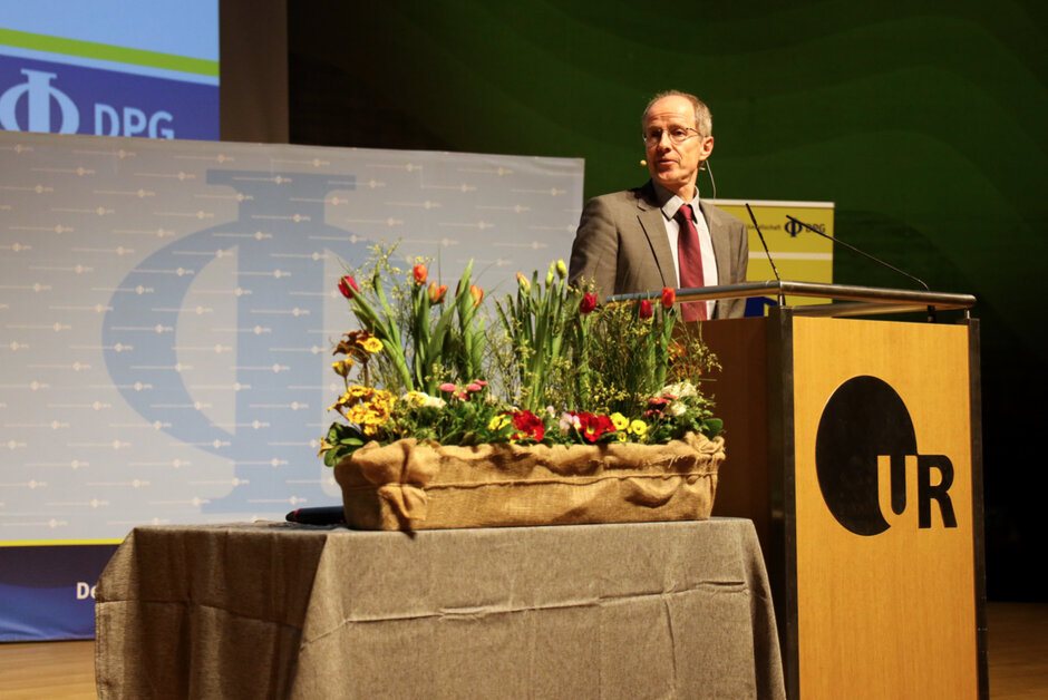 Ein Mann im Anzug, mit Brille und Kopfmikrophon steht hinter einem mit Uni-Regensburg-Logo geschmückten Rednerpult.