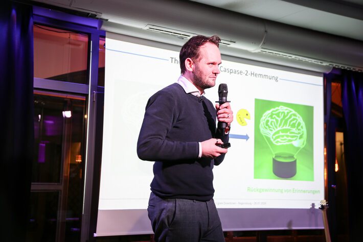 Eine Person steht auf einer Bühne vor einer hellen Präsentationsfolie und hält ein Mikrofon in der Hand. Auf der Folie sind ein Pfeildiagramm sowie eine Illustration eines Gehirns zu sehen. Die Szene wirkt wie ein wissenschaftlicher Vortrag oder eine Präsentation in einem Veranstaltungsraum. (öffnet Vergrößerung des Bildes)