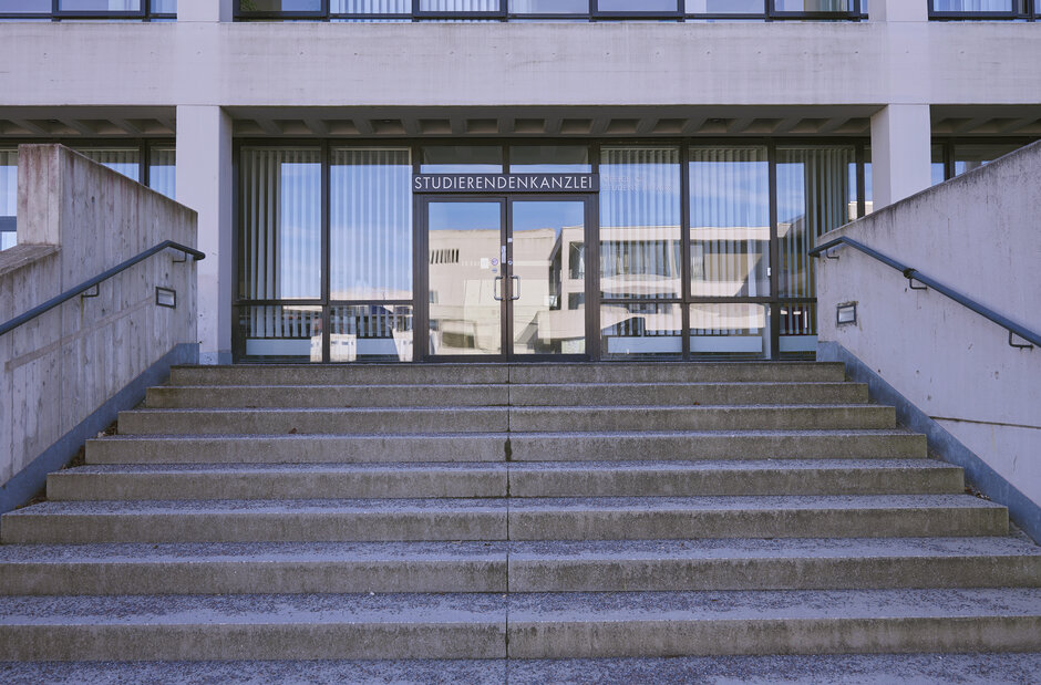 Eine breite Treppe führt zu einem Gebäude mit Fensterfront und Eingangstür, über der eine Beschriftung mit "Studierendenkanzlei" angebracht ist.