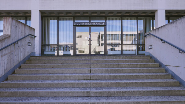 Eine breite Treppe führt zu einem Gebäude mit Fensterfront und Eingangstür, über der eine Beschriftung mit "Studierendenkanzlei" angebracht ist.