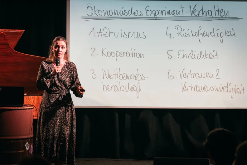 Eine Frau steht auf einer Bühne und spricht in ein Mikrofon, das sie in der rechten Hand hält. In ihrer linken Hand hält sie einen Presenter. Im Hintergrund ist auf einer Leinwand ein handschriftlicher Text projeziert: "Ökonomisches Experiment-Verhalten. 1. Altruismus 2. Kooperation 3. Wettbewerbsbereitschaft 4. Risikofreudigkeit 5. Ehrlichkeit 6. Vertrauen & Vertrauenswürdigkeit (öffnet Vergrößerung des Bildes)
