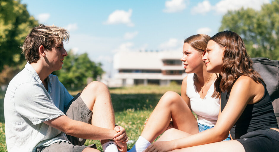 Studierende sitzen zusammen auf einer Picknickdecke auf einer Wiese am Campus der Universität Regensburg.