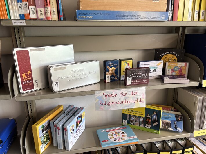 Auf einem Regalbrett liegt eine Auswahl verschiedener Boxen mit Spielen für den Religionsunterricht (öffnet Vergrößerung des Bildes)