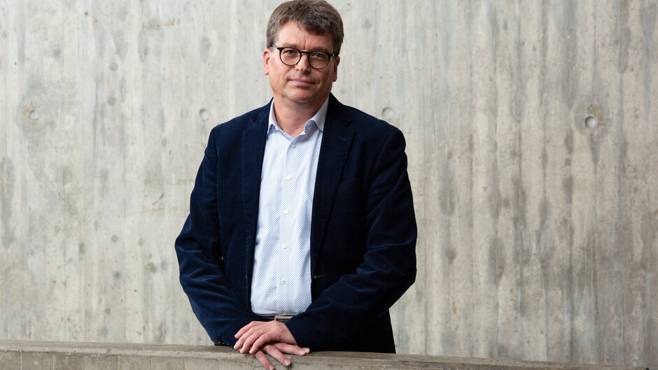 Mann mit Brille, in Jacket und Hemd, stehend vor Betonwand.