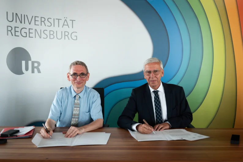 Zwei Männer sitzen an einem Konferenztisch. Sie halten Stifte in der jeweils rechten Hand und sind im Begriff, vor ihnen liegende Papiere zu unterschreiben. Im Hintergrund ist ein vielfarbiges Banner mit dem Schrift „Universität Regensburg“ zu sehen. (öffnet Vergrößerung des Bildes)