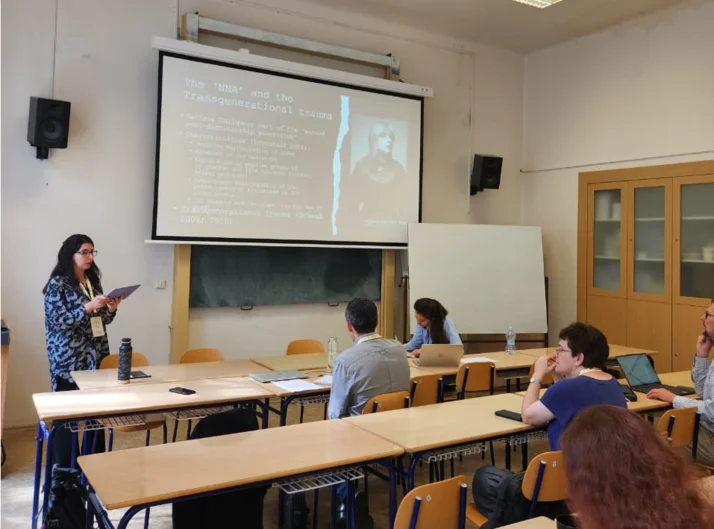 Bárbara Aranda stands at the lectern in a seminar room. Students sit at tables.