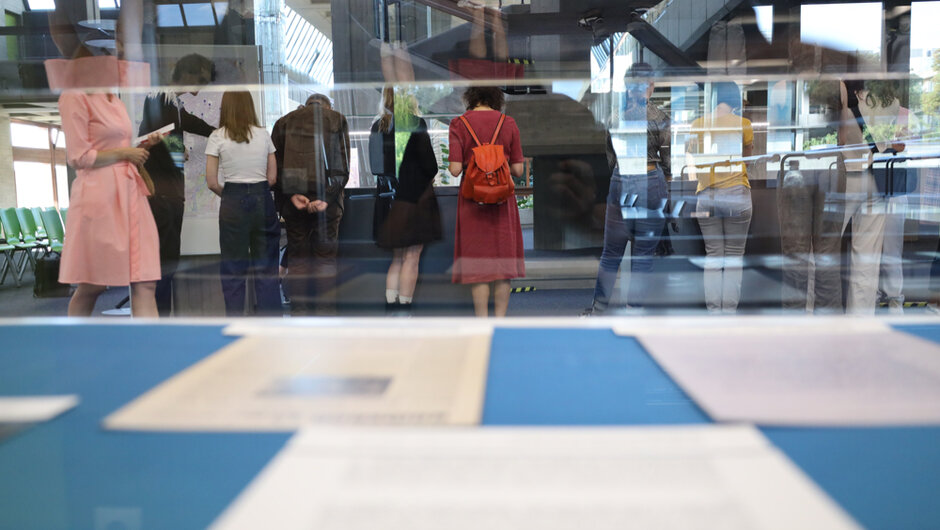 Durch eine Glasvitrine hindurch geht der Blick auf 10 Personen, die sich über Exponate in Vitrinen beugen. Sie sind von hinten zu sehen. 