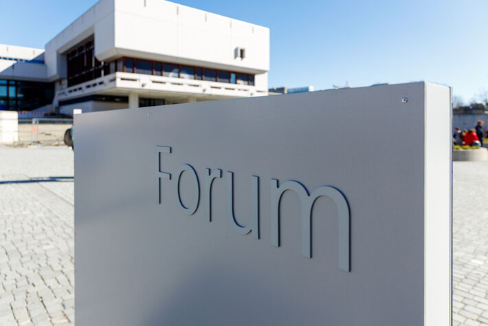 Schild, auf dem Forum steht, im Hintergrund die Zentralbibliothek der Uni Regensburg (öffnet Vergrößerung des Bildes)