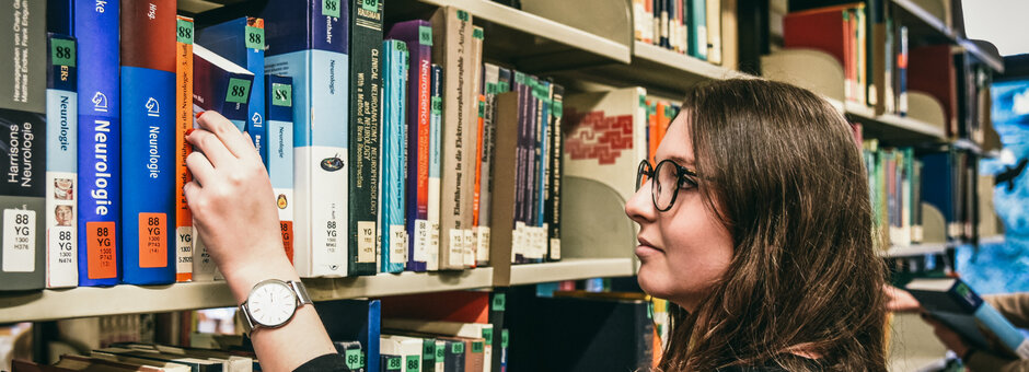 Eine junge Frau wählt an einem Bücherregal im Lesesaal Biologie der Universität Regensburg ein Buch aus.