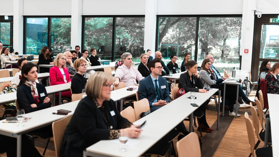 In a lecture hall, several people are sitting at tables. Their attention is focused on the podium.