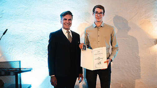 Zwei Herren stehen vor einer weißen Wand, der rechts stehende trägt ein beiges Hemd und hält eine aufgeschlagene Urkundenmappe in der Hand. Er hält sie so, dass die Betrachterinnen und Betrachter die enthaltene Urkunde sehen können. 