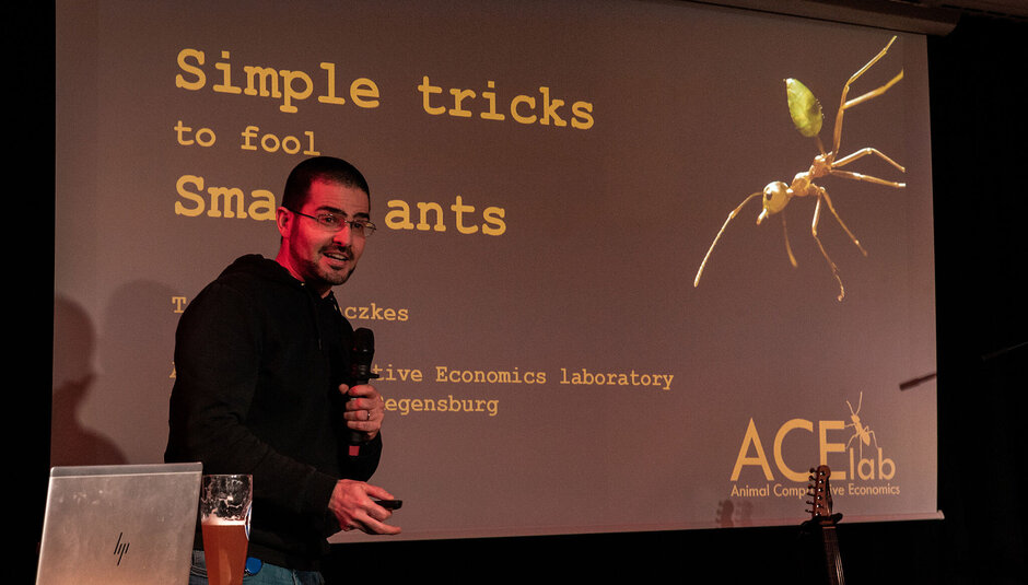 Ein Mann mit einem Mikrofon in der einen und einer Art Fernbedienung in der anderen Hand steht vor einer Beamer-Projektion und spricht.