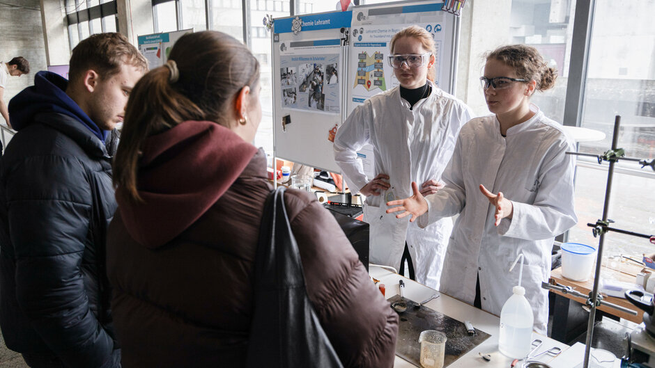 Junger Mann und junge Frau in Laborkitteln an Labortisch, davor stehen andere junge Menschen. (öffnet Vergrößerung des Bildes)