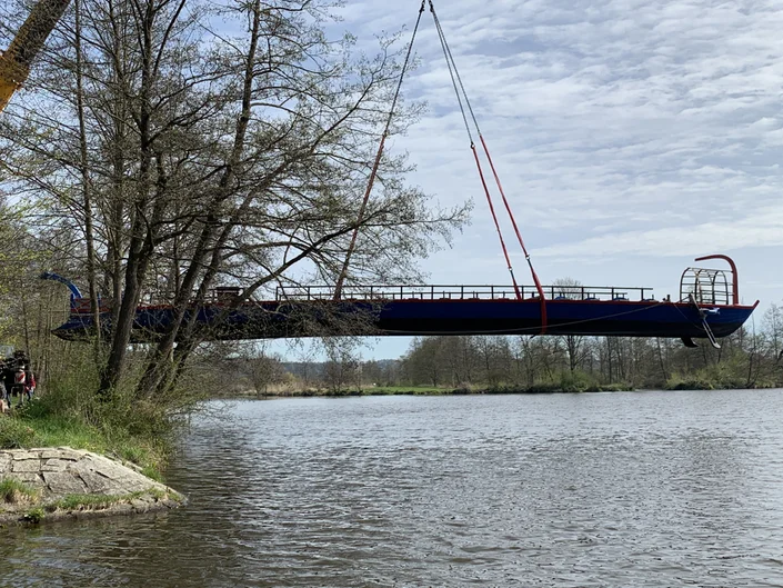 Ein Schiff schwebt, von roten Gurten gehalten, einige Meter über einer Wasseroberfläche. Das Ufer ist befestigt und mit Büschen und Bäumen bewachsen. Die Bäume sind spärlich belaubt, es scheint Frühling zu sein.