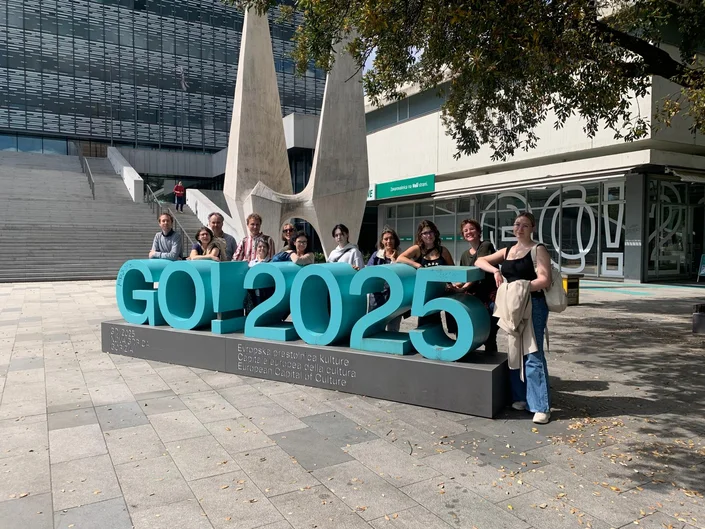 Die Exkursionsgruppe steht auf einem Platz um einen etwa 3 Meter breiten, plastischen Schriftzug "Go! 2025" in türkiser Farbe herum. Im Hintergrund sind Hochhausfassaden erkennbar.  (öffnet Vergrößerung des Bildes)