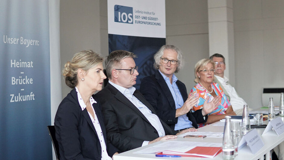 Fünf Personen sitzen an einem langen Tisch, auf dem Flaschen, Gläser und Namenssteller stehen sowie Unterlagen und Smartphones liegen. Der Mann in der Mitte spricht gestikulierend. Im Hintergrund steht ein Roll-Up mit der Aufschrift "IOS". (öffnet Vergrößerung des Bildes)