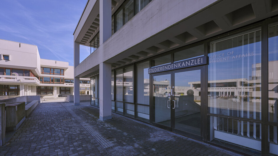 Der Eingang in die Studierendenkanzlei der Universität Regensburg; in den Glasscheiben der Fenster und der Eingangstür spiegelt sich das Forum der Universität.