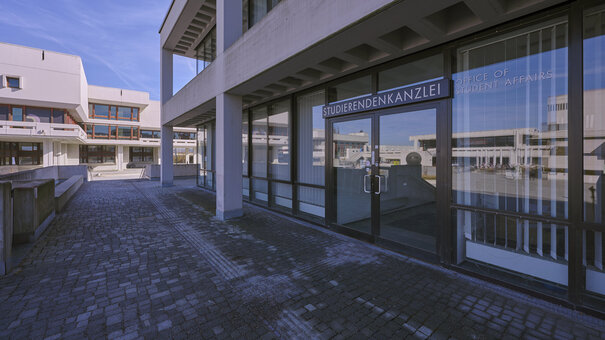 Der Eingang in die Studierendenkanzlei der Universität Regensburg; in den Glasscheiben der Fenster und der Eingangstür spiegelt sich das Forum der Universität.