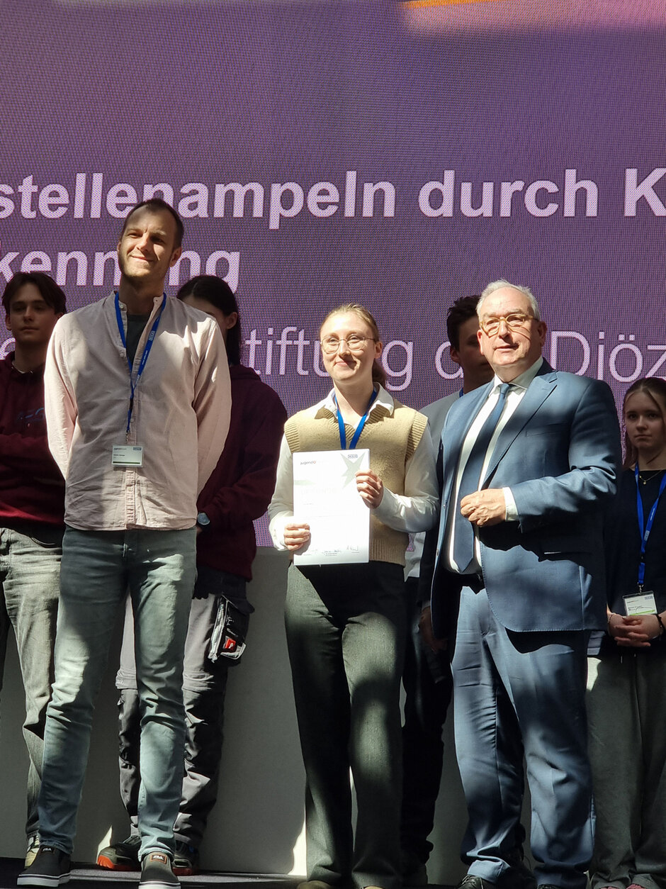 Mehrere Menschen stehen auf einer Bühne, im Sonnenschein. Die junge Frau in der Mitte hält lächelnd eine Urkunde in den Händen.