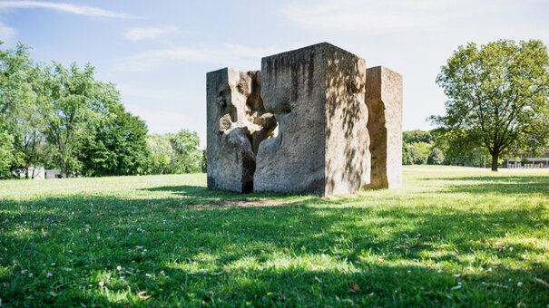 Ein Kunstwerk aus Granit, das einem auseinandergebrochenen Würfel ähnelt und auf einer sonnenbeschienenen, grünen Wiese steht. 