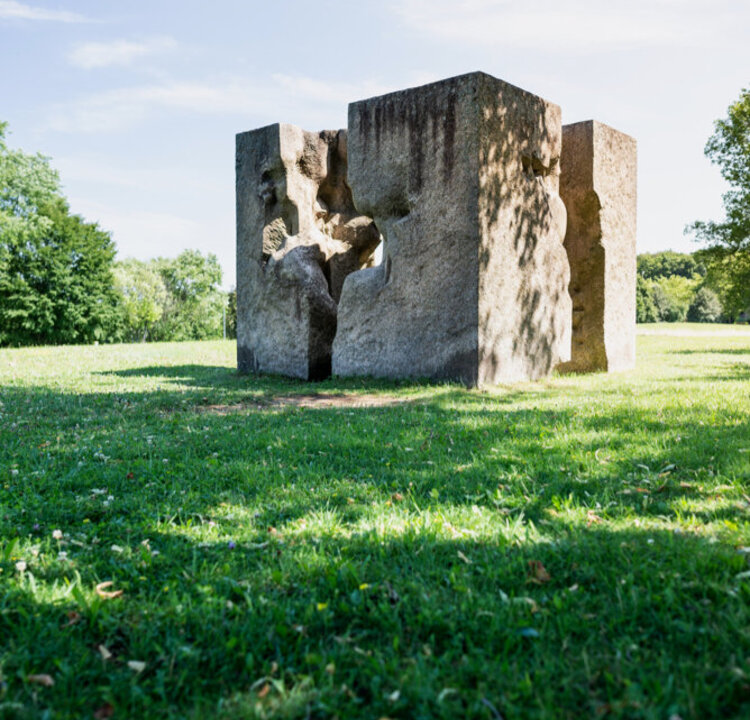 Ein Kunstwerk aus Granit, das einem auseinandergebrochenen Würfel ähnelt und auf einer sonnenbeschienenen, grünen Wiese steht. 