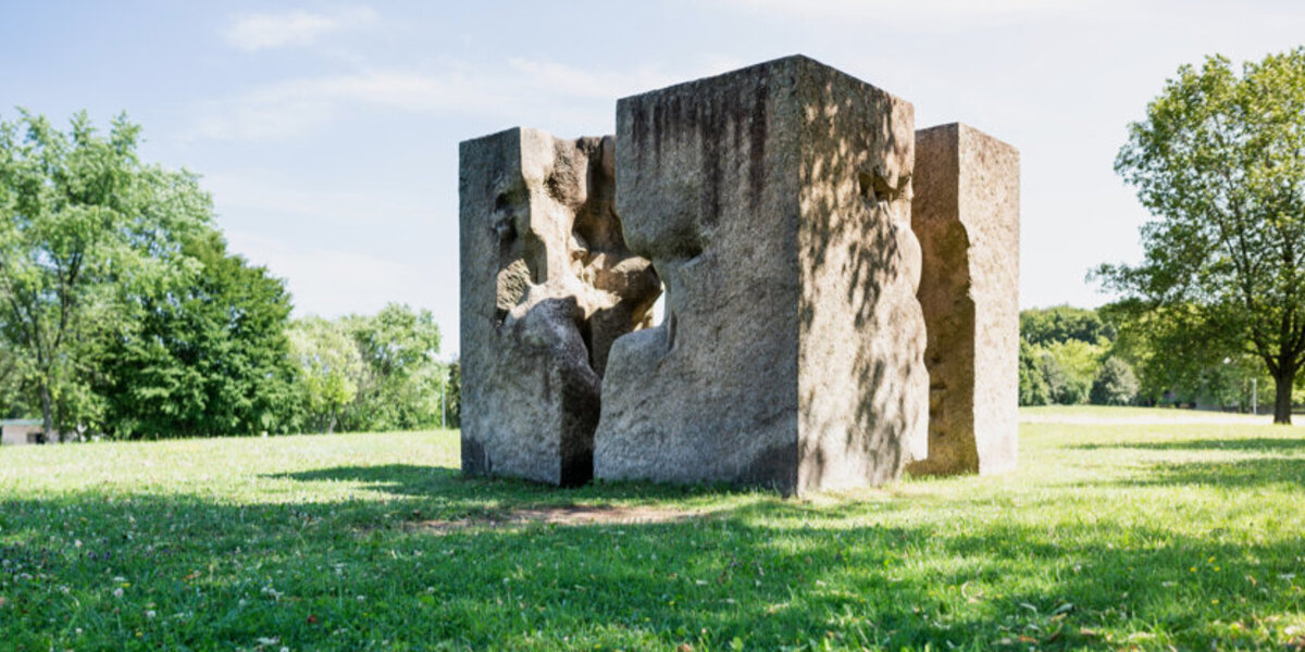 Ein Kunstwerk aus Granit, das einem auseinandergebrochenen Würfel ähnelt und auf einer sonnenbeschienenen, grünen Wiese steht. 