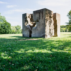 Ein Kunstwerk aus Granit, das einem auseinandergebrochenen Würfel ähnelt und auf einer sonnenbeschienenen, grünen Wiese steht. 