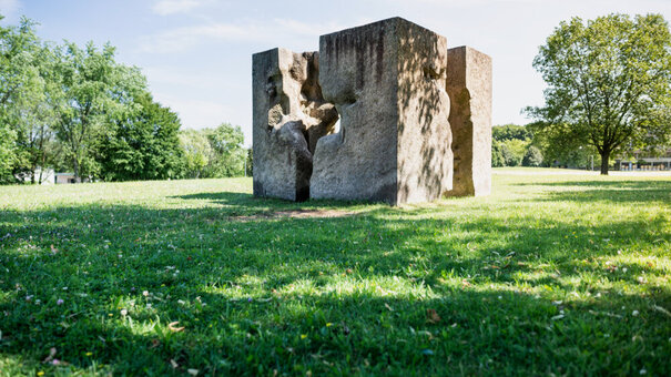 Ein Kunstwerk aus Granit, das einem auseinandergebrochenen Würfel ähnelt und auf einer sonnenbeschienenen, grünen Wiese steht. 