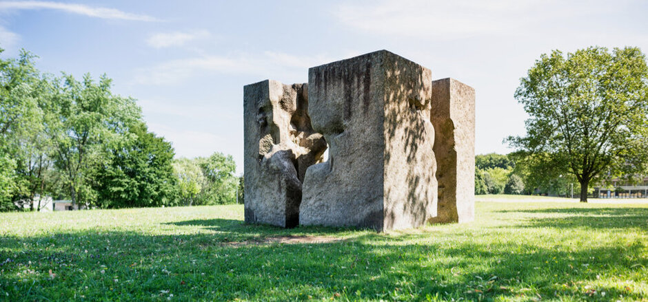 Ein Kunstwerk aus Granit, das einem auseinandergebrochenen Würfel ähnelt und auf einer sonnenbeschienenen, grünen Wiese steht. 