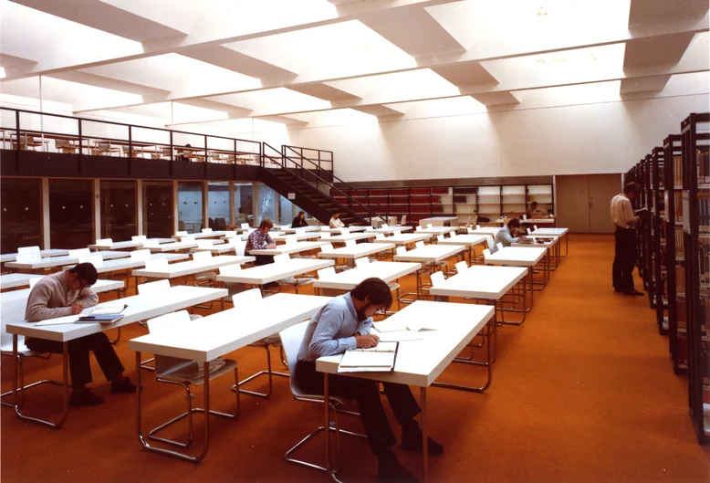 In einem Lesesaal mit orange-farbenem Teppichboden stehen weiße Tische und Stühle, an den Wänden befinden sich Bücherregale. An einigen Tischen sitzen junge Personen; sie lesen oder schreiben. Die Farbgebung des Fotos weist auf seinen Ursprung in den 1970er Jahren hin. (öffnet Vergrößerung des Bildes)