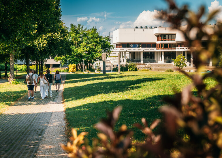 Eine Gruppe junger Leute geht an einem Sommerabend über den grünen Campus der Universität Regensburg. Im Bildhintergrund ist die Zentralbibliothek zu sehen.