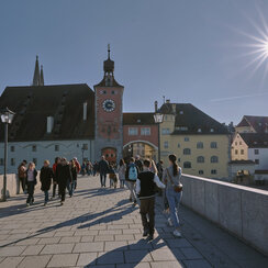 Passanten schlendern bei Sonnenschein über eine mittelalterlicher Steinbrücke. Im Hintergrund bunte, historiche Häuserfassaden.