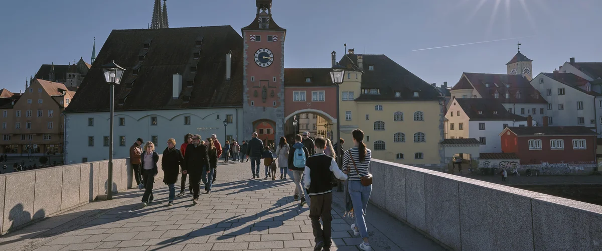 Passanten schlendern bei Sonnenschein über eine mittelalterlicher Steinbrücke. Im Hintergrund bunte, historiche Häuserfassaden.