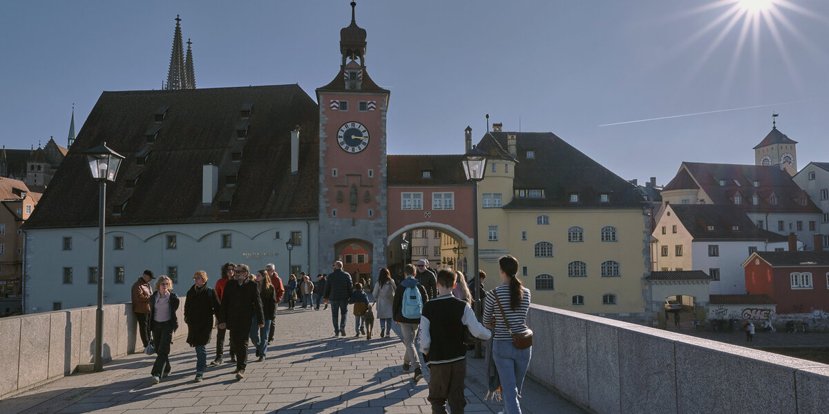 Passanten schlendern bei Sonnenschein über eine mittelalterlicher Steinbrücke. Im Hintergrund bunte, historiche Häuserfassaden.
