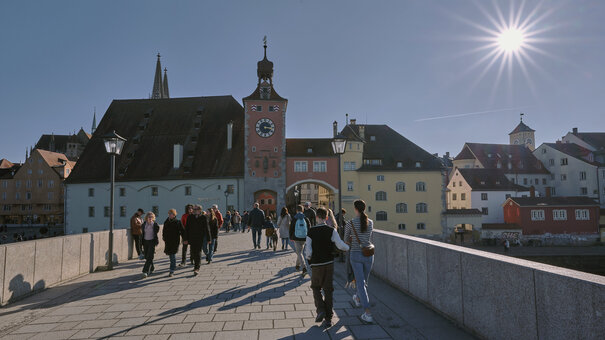 Passanten schlendern bei Sonnenschein über eine mittelalterlicher Steinbrücke. Im Hintergrund bunte, historiche Häuserfassaden.