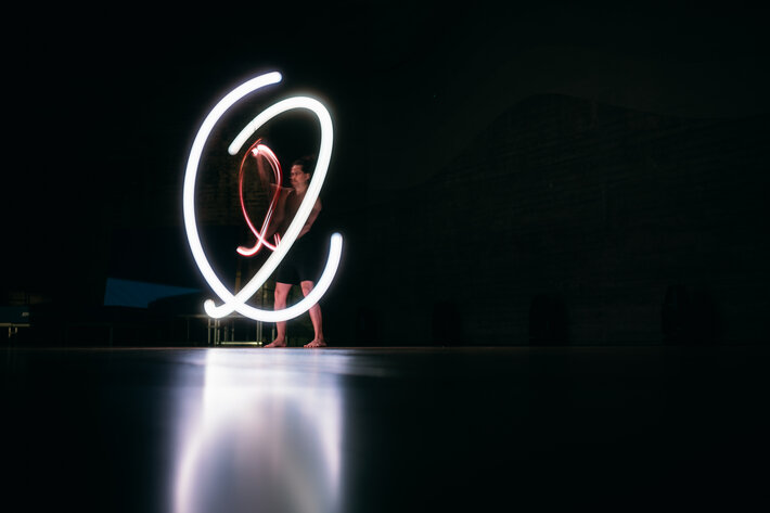 Ein Akrobat steht im Dunkeln. Zu sehen sind die Linien, die er mit einem leuchtenden Gegenstand in die Luft zeichnet. (öffnet Vergrößerung des Bildes)