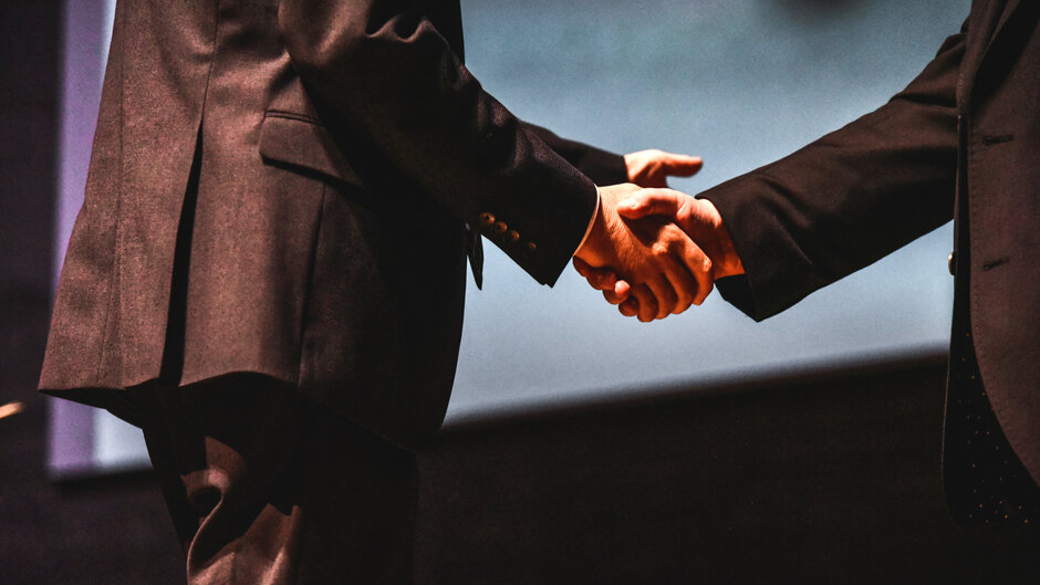 Zwei Personen in dunklen Anzügen schütteln sich die Hände.