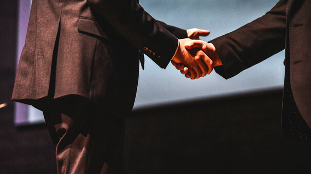 Zwei Personen in dunklen Anzügen schütteln sich die Hände.