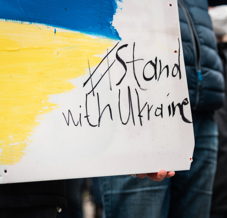 Bei einer Demonstration wird ein Plakat mit gelber und blauer Farbe sowie dem Schriftzug "Stand with Ukraine" gezeigt.