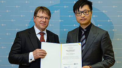 Zwei Männer in formeller Kleidung halten eine geöffnete Dokumentenmappe mit einem formellen Schreiben darin in Richtung Betrachter.