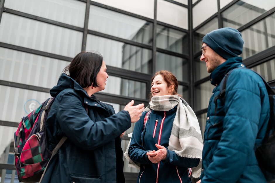 Drei Personen stehen am Campus der Universität Regensburg zusammen und unterhalten sich.