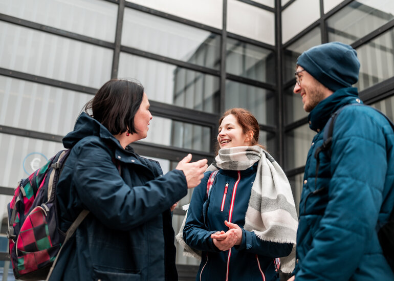 Drei Personen stehen am Campus der Universität Regensburg zusammen und unterhalten sich.